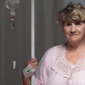 Donna anziana in camicia da notte riceve una flebo in un contesto domestico o ospedaliero, con sguardo tranquillo.
