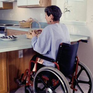 Donna in sedia a rotelle prepara del cibo in una cucina accessibile, con utensili e fornelli adattati al piano ribassato.