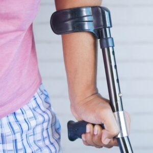 Dettaglio del braccio di una persona che impugna una stampella ergonomica nera, indossa un pigiama a righe e una maglietta rosa.